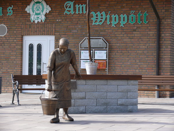 Bronzefigur einer alten Frau mit Wassereimer vor einem Ziehbrunnen